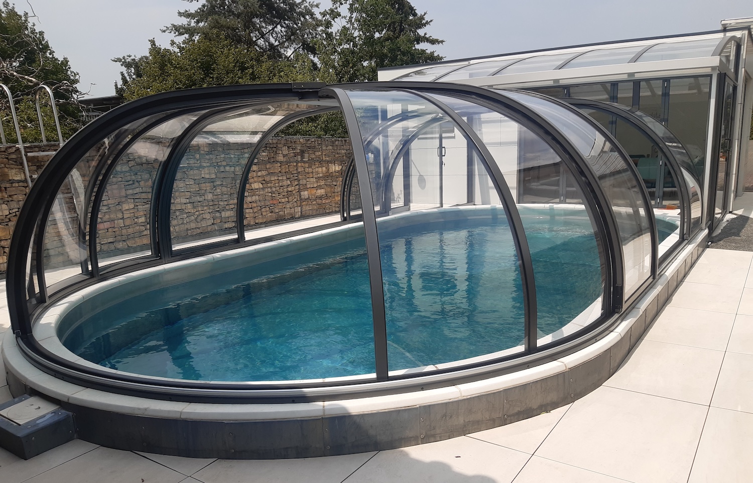 Halbhohe, transparente Poolüberdachung über einem ovalen Außenpool mit Verbindung zu einer Terrasse und einem Wohnhaus.