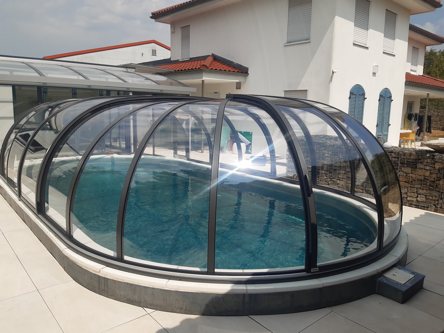 Halbhohe, transparente Poolüberdachung über einem ovalen Außenpool mit Verbindung zur Terrasse und zum Wohnhaus.
