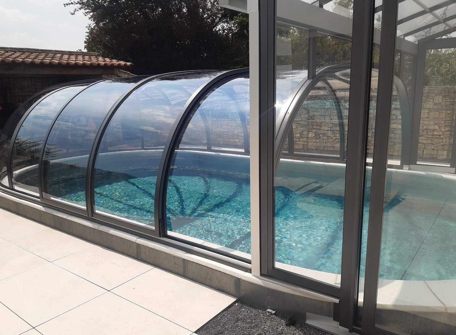 Seitliche Ansicht einer geschlossenen Poolüberdachung mit gebogenen Glaselementen und Verbindung zur Terrasse.