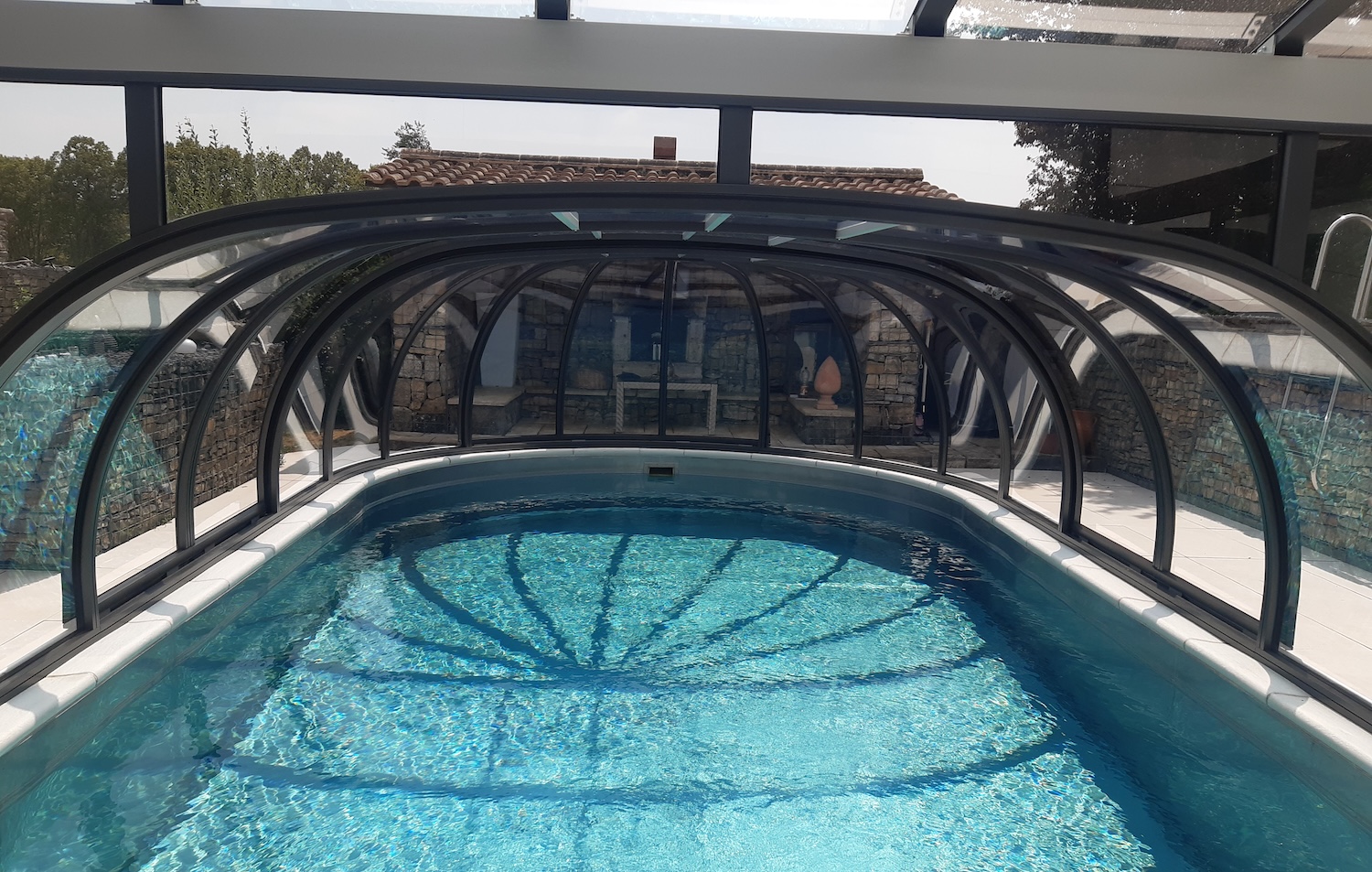 Blick unter eine transparente Poolüberdachung auf einen geschützten Außenpool mit klarem Wasser.