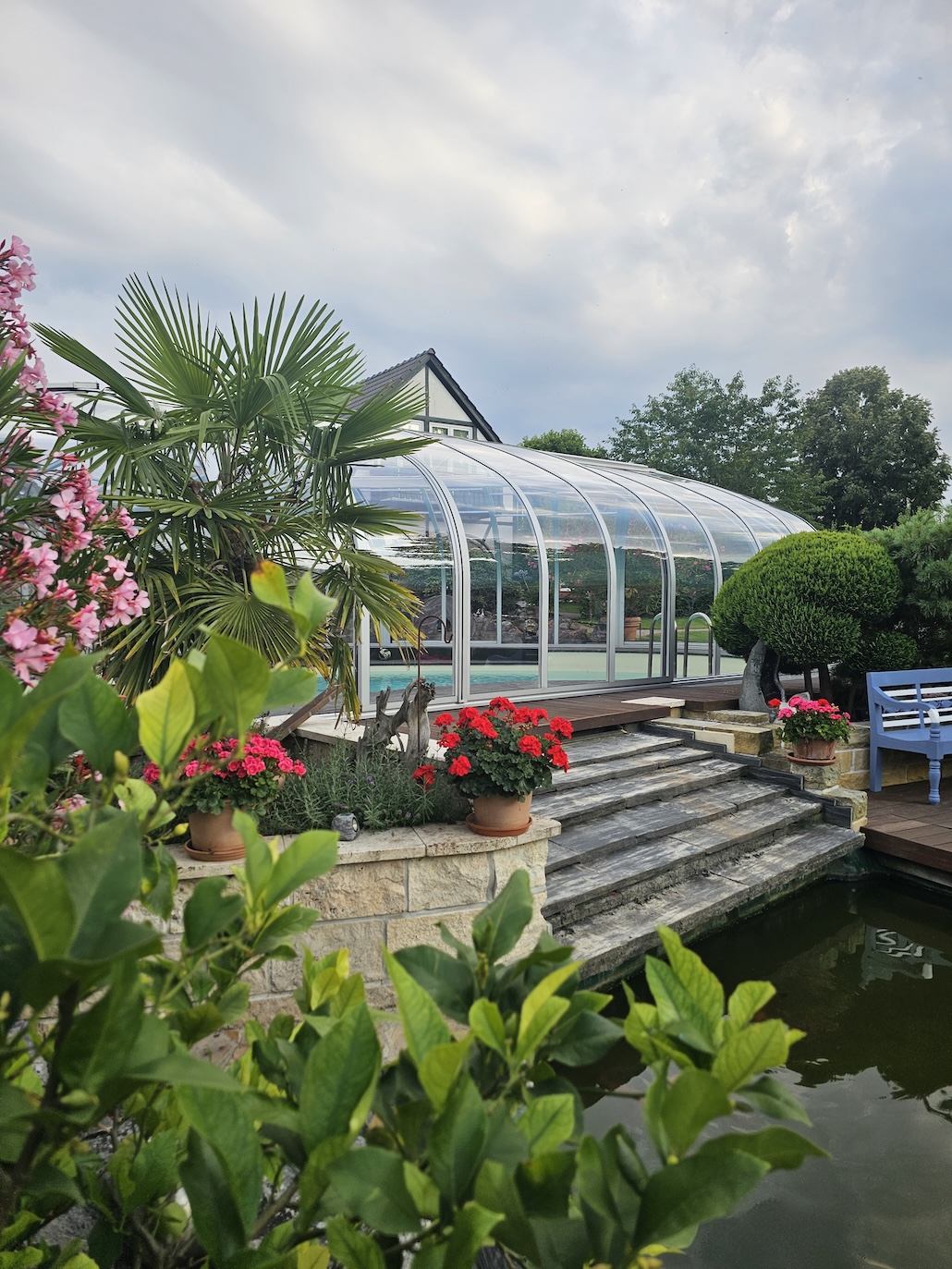 Transparente Poolüberdachung in einem üppig bepflanzten Garten mit Palmen, Blumen und Steintreppe