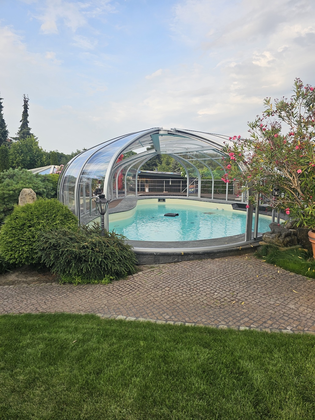 Hohe transparente Poolüberdachung über einem geschwungenen Außenpool, umgeben von Sträuchern und Blumen